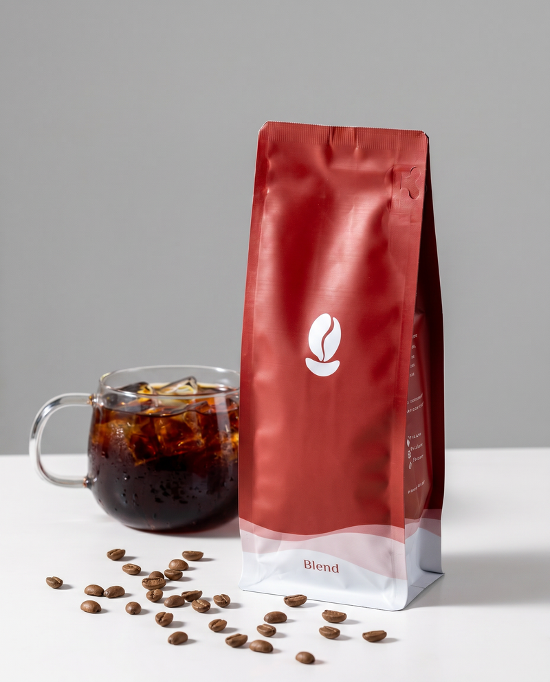 A red Bean Shipper Blend bag beside a glass mug of iced black coffee with coffee beans on a white table