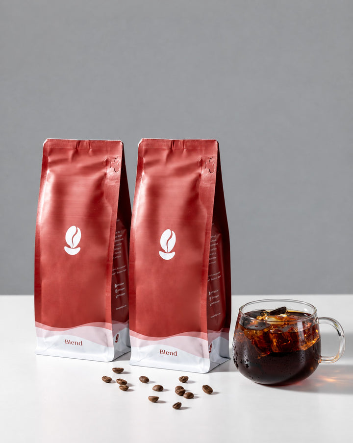 Bestseller Bundle: two 300g Bean Shipper bags and a glass of iced black coffee on a white table against a grey wall