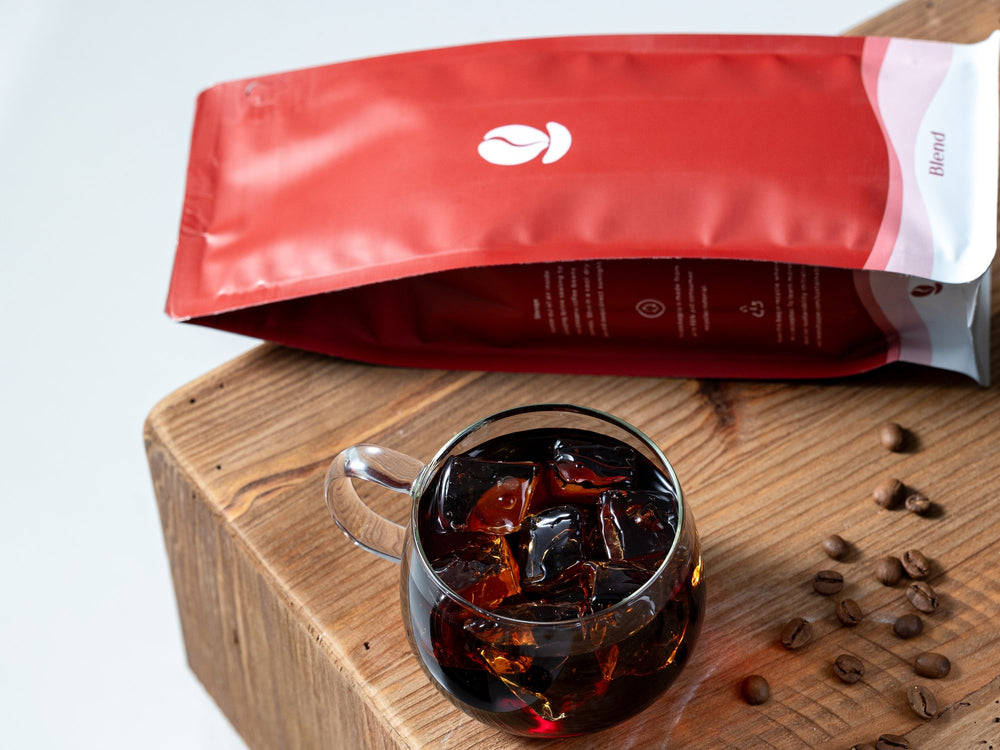 A red Bean Shipper Blend bag lying on a wooden block beside a glass of iced coffee